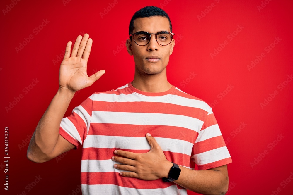 Young handsome african american man wearing casual striped t-shirt and glasses Swearing with hand on chest and open palm, making a loyalty promise oath Young handsome african american man wearing casual striped t-shirt and glasses Swearing with hand on chest and open palm, making a loyalty promise oath