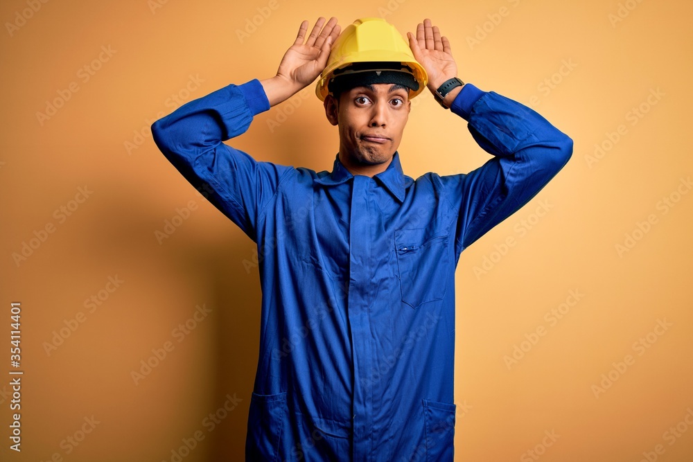 Young handsome african american worker man wearing blue uniform and security helmet Doing bunny ears gesture with hands palms looking cynical and skeptical. Easter rabbit concept.