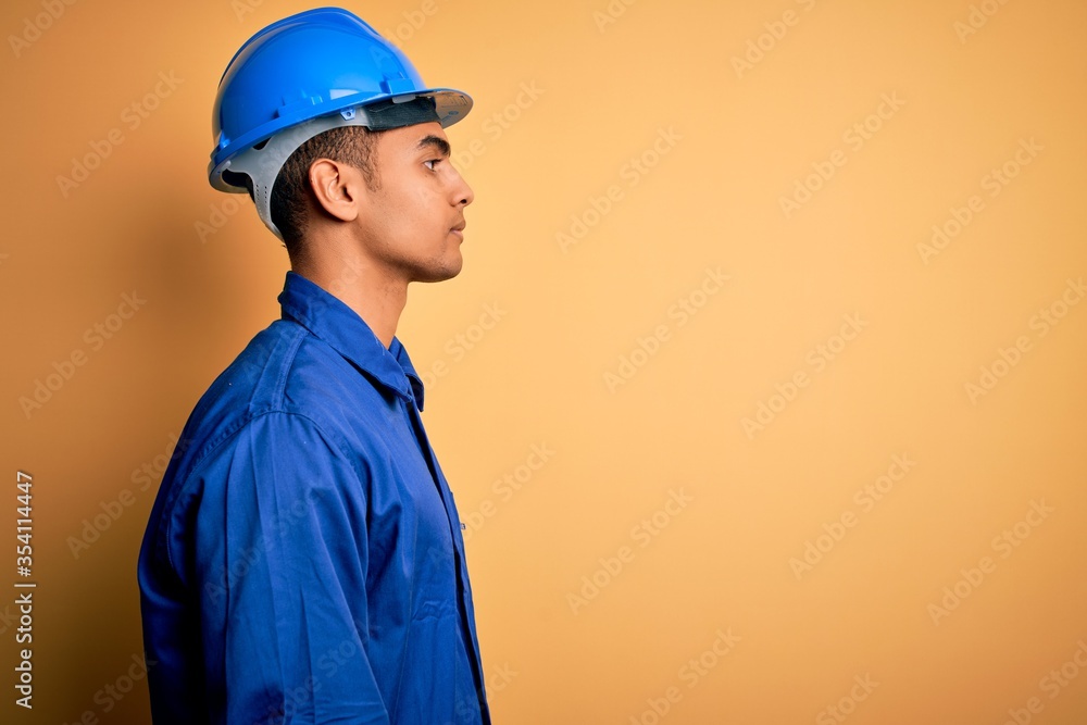 Young handsome african american worker man wearing blue uniform and security helmet looking to side, relax profile pose with natural face with confident smile.