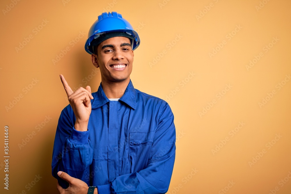 Young handsome african american worker man wearing blue uniform and security helmet with a big smile on face, pointing with hand and finger to the side looking at the camera.