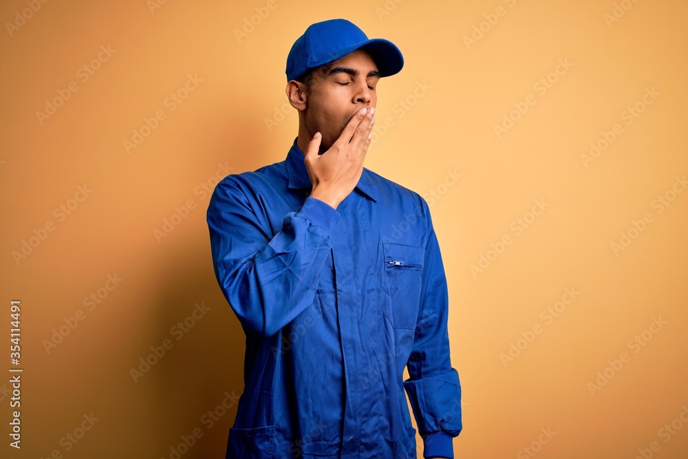Young african american mechanic man wearing blue uniform and cap over yellow background bored yawning tired covering mouth with hand. Restless and sleepiness.