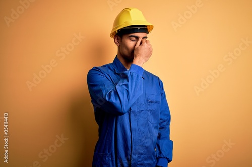 Young handsome african american worker man wearing blue uniform and security helmet tired rubbing nose and eyes feeling fatigue and headache. Stress and frustration concept.