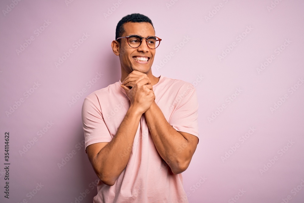 Handsome african american man wearing casual t-shirt and glasses over pink background laughing nervous and excited with hands on chin looking to the side Handsome african american man wearing casual t-shirt and glasses over pink background laughing nervous and excited with hands on chin looking to the side