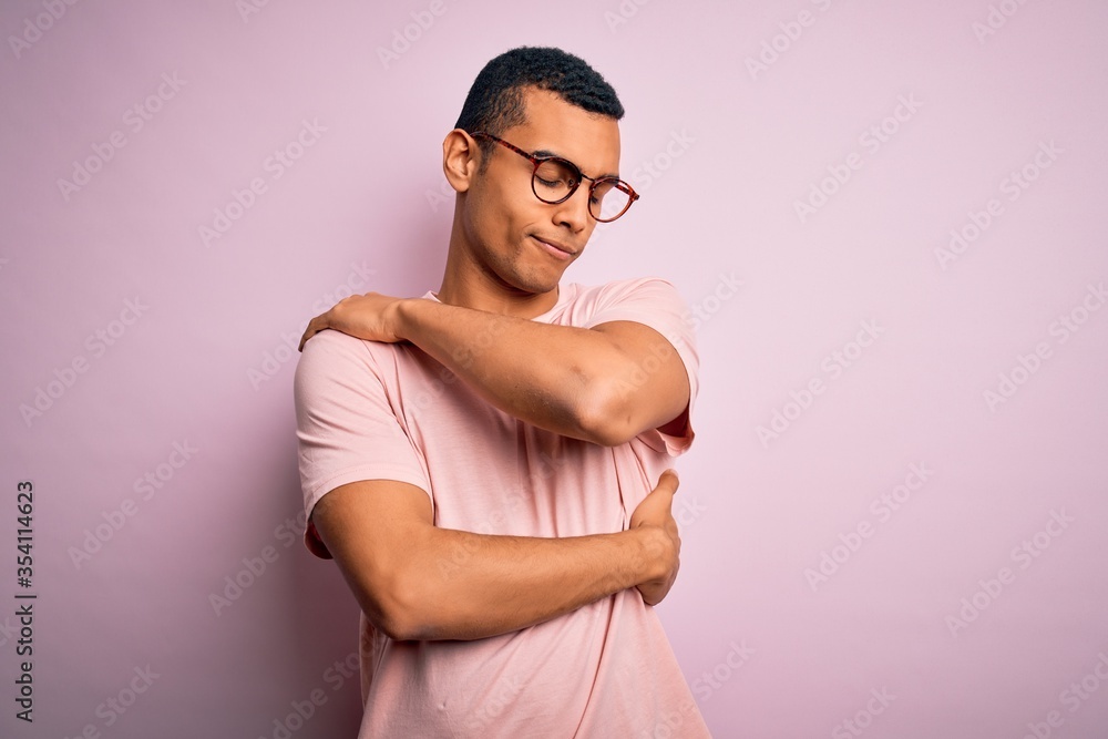 Obraz premium Handsome african american man wearing casual t-shirt and glasses over pink background Hugging oneself happy and positive, smiling confident. Self love and self care