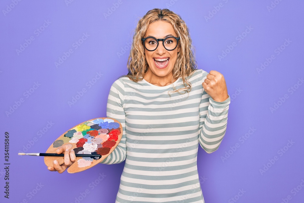 Young beautiful blonde artist woman drawing using paintbrush and palette with colors screaming proud, celebrating victory and success very excited with raised arms Young beautiful blonde artist woman drawing using paintbrush and palette with colors screaming proud, celebrating victory and success very excited with raised arms