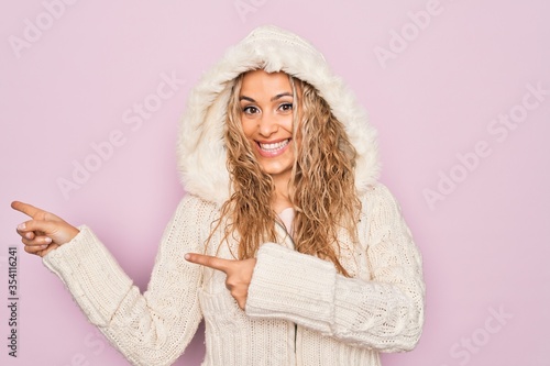 Young beautiful blonde woman wearing casual sweater with hood over isolated pink background smiling and looking at the camera pointing with two hands and fingers to the side.