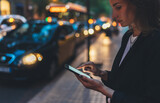 Elegant woman pointing on screen smartphone background headlights auto in night city street, tourist girl using internet technology calls a taxi standing next to the road in busy street of evening
