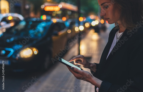 Elegant woman pointing on screen smartphone background headlights auto in night city street, tourist girl using internet technology calls a taxi standing next to the road in busy street of evening photo