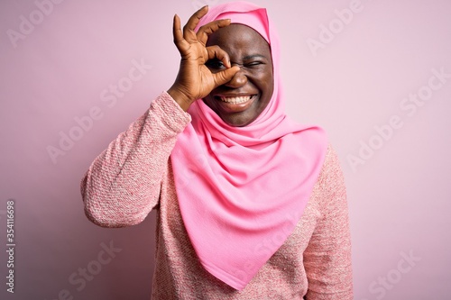 Young african american plus size woman wearing muslim hijab over isolated pink background doing ok gesture with hand smiling, eye looking through fingers with happy face.