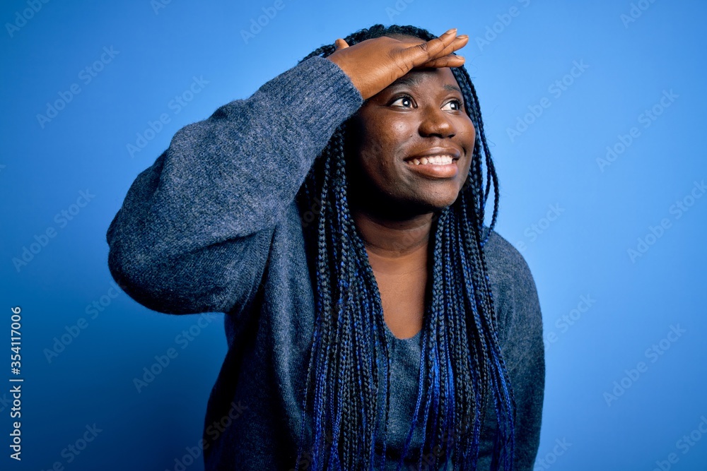 African american plus size woman with braids wearing casual sweater over blue background very happy and smiling looking far away with hand over head. Searching concept. African american plus size woman with braids wearing casual sweater over blue background very happy and smiling looking far away with hand over head. Searching concept.