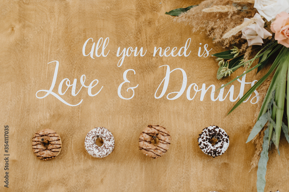 Fototapeta premium Wooden board with various glazed donuts and powder. Caption: all we need is love and donuts. Arrangement of flowers and tropical leaves