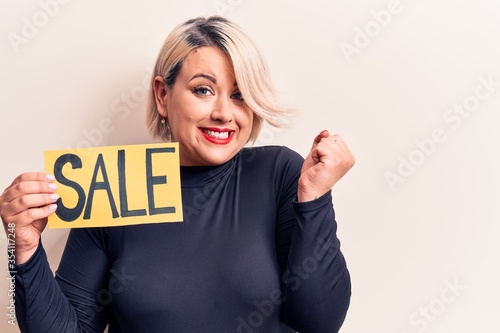 Young beautiful blonde plus size woman holding sale banner over isolated white background screaming proud, celebrating victory and success very excited with raised arm