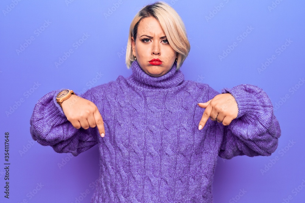 Obraz premium Beautiful blonde plus size woman wearing casual turtleneck sweater over purple background Pointing down looking sad and upset, indicating direction with fingers, unhappy and depressed.