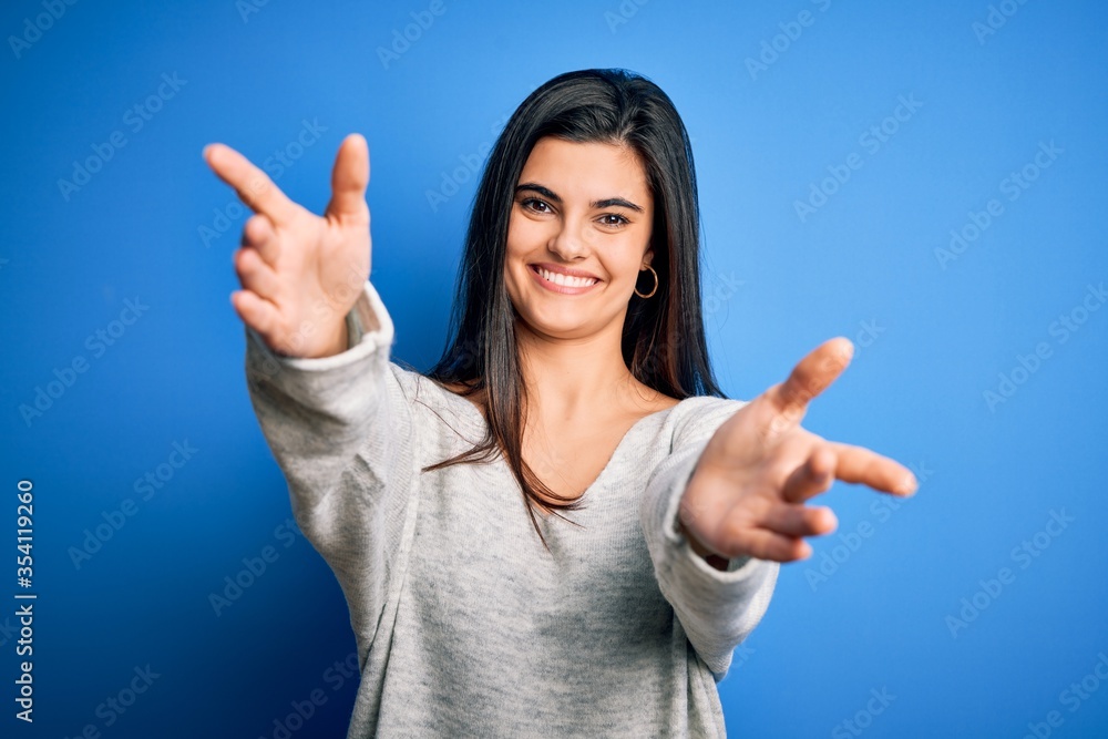 Young beautiful brunette woman wearing casual sweater standing over blue background looking at the camera smiling with open arms for hug. Cheerful expression embracing happiness. Young beautiful brunette woman wearing casual sweater standing over blue background looking at the camera smiling with open arms for hug. Cheerful expression embracing happiness.