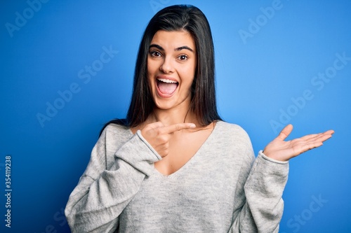 Young beautiful brunette woman wearing casual sweater standing over blue background amazed and smiling to the camera while presenting with hand and pointing with finger.