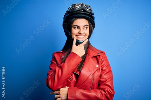 Young beautiful brunette motorcycliste woman wearing motorcycle helmet and jacket with hand on chin thinking about question, pensive expression. Smiling with thoughtful face. Doubt concept. © Krakenimages.com