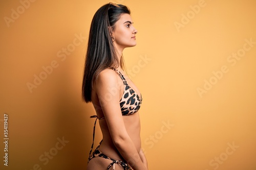 Young beautiful brunette woman on vacation wearing swimwear bikini over yellow background looking to side, relax profile pose with natural face with confident smile.