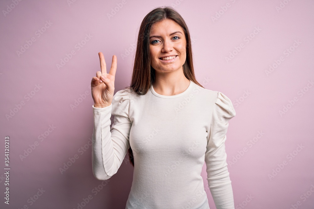 Young beautiful woman with blue eyes wearing casual white t-shirt over pink background smiling looking to the camera showing fingers doing victory sign. Number two. Young beautiful woman with blue eyes wearing casual white t-shirt over pink background smiling looking to the camera showing fingers doing victory sign. Number two.