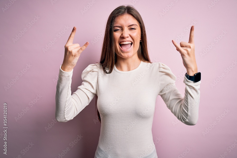 Young beautiful woman with blue eyes wearing casual white t-shirt over pink background shouting with crazy expression doing rock symbol with hands up. Music star. Heavy music concept. Young beautiful woman with blue eyes wearing casual white t-shirt over pink background shouting with crazy expression doing rock symbol with hands up. Music star. Heavy music concept.