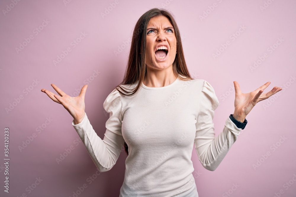 Young beautiful woman with blue eyes wearing casual white t-shirt over pink background crazy and mad shouting and yelling with aggressive expression and arms raised. Frustration concept. Young beautiful woman with blue eyes wearing casual white t-shirt over pink background crazy and mad shouting and yelling with aggressive expression and arms raised. Frustration concept.