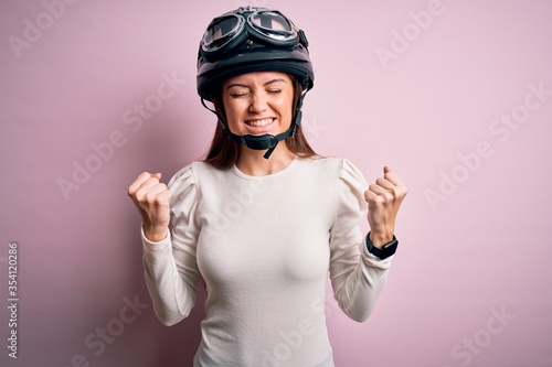 Young beautiful motorcyclist woman with blue eyes wearing moto helmet over pink background very happy and excited doing winner gesture with arms raised, smiling and screaming for success. Celebration