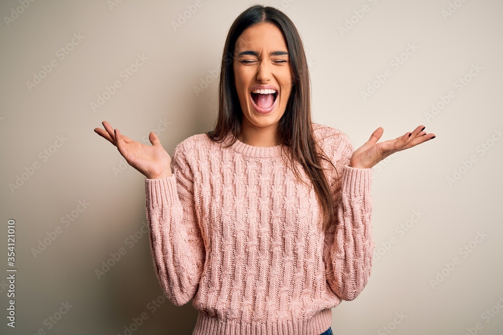 Young beautiful brunette woman wearing casual sweater over isolated white background celebrating mad and crazy for success with arms raised and closed eyes screaming excited. Winner concept Young beautiful brunette woman wearing casual sweater over isolated white background celebrating mad and crazy for success with arms raised and closed eyes screaming excited. Winner concept