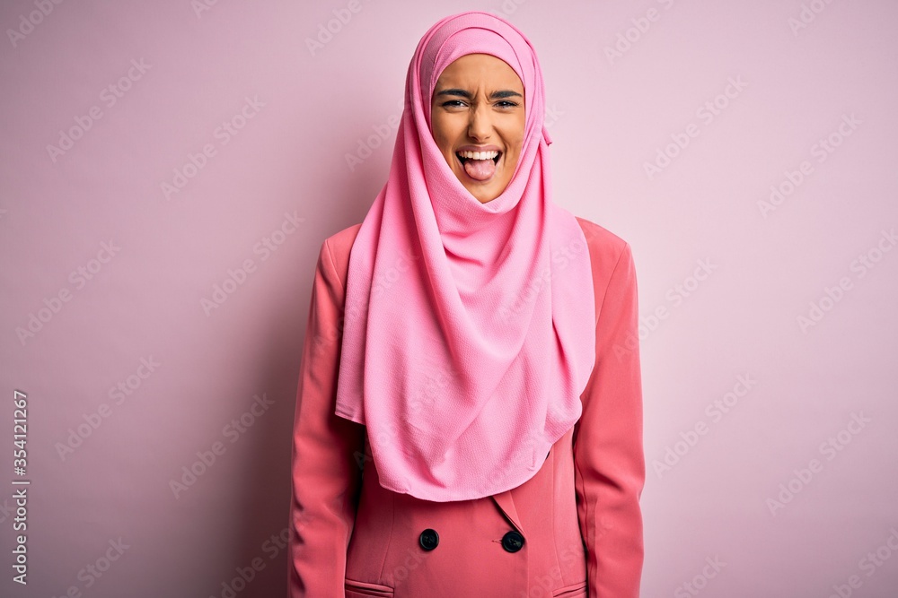 Fototapeta premium Young beautiful brunette businesswoman wearing pink muslim hijab and business jacket sticking tongue out happy with funny expression. Emotion concept.