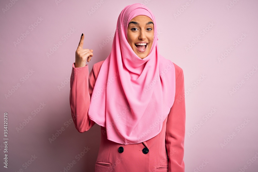 Young beautiful brunette businesswoman wearing pink muslim hijab and business jacket pointing finger up with successful idea. Exited and happy. Number one. Young beautiful brunette businesswoman wearing pink muslim hijab and business jacket pointing finger up with successful idea. Exited and happy. Number one.