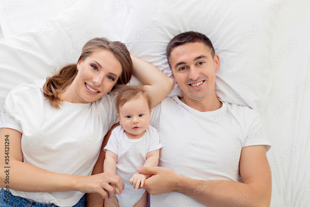 Happily family: mother, father and son, a boy, lie on their backs on a white bed in a sunny bedroom. Parent and little child are resting at home. View from above