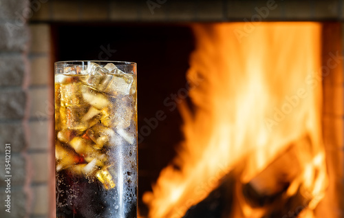 Glass of cola with ice in front of burning fireplace in a country house.