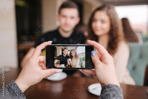 Hand of female making photo of beutiful young couple in cafe. Happy smiled couple posing for photo on smartphone