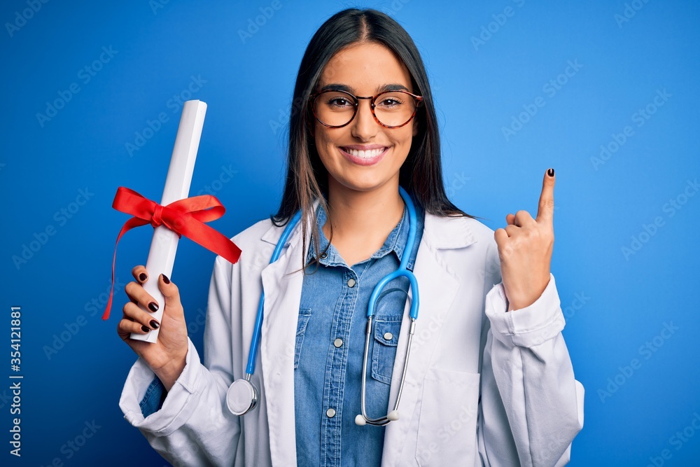 Young beautiful brunette doctor woman wearing glasses and coat holding diploma degree surprised with an idea or question pointing finger with happy face, number one Young beautiful brunette doctor woman wearing glasses and coat holding diploma degree surprised with an idea or question pointing finger with happy face, number one