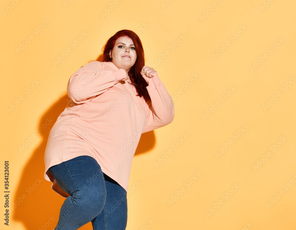 Concentrated fatty young female in jeans and sweater posing in fighting stance preparing to kick. Studio shot isolated on orange, copy space Concentrated fatty young female in jeans and sweater posing in fighting stance preparing to kick. Studio shot isolated on orange, copy space