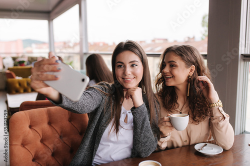 Attractive young women making selfie in restairant. Women drin cappuccino on terrace. Two female make photo together photo