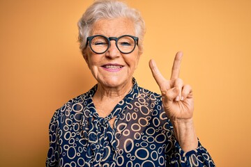 Senior beautiful grey-haired woman wearing casual shirt and glasses over yellow background smiling with happy face winking at the camera doing victory sign. Number two.