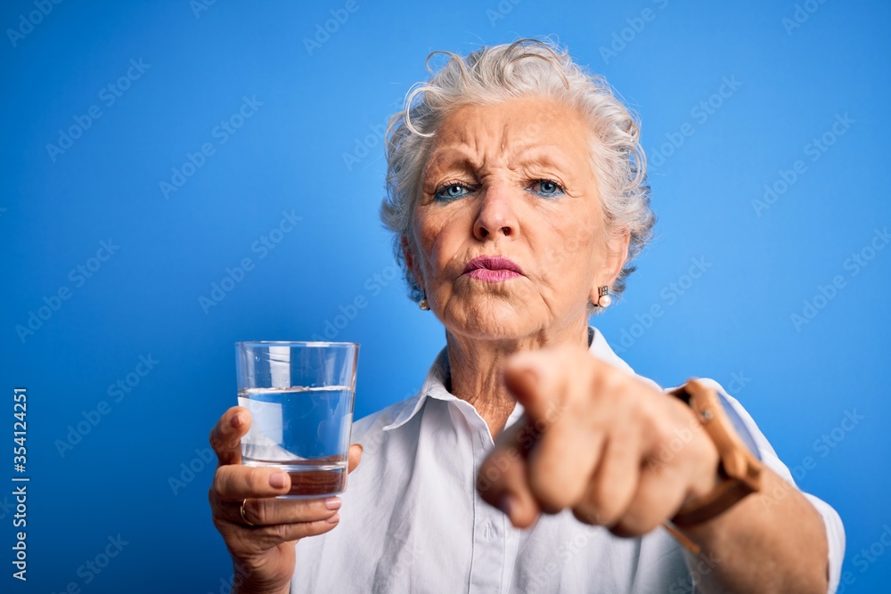 Senior beautiful woman drinking glass of water standing over isolated blue background pointing with finger to the camera and to you, hand sign, positive and confident gesture from the front Senior beautiful woman drinking glass of water standing over isolated blue background pointing with finger to the camera and to you, hand sign, positive and confident gesture from the front