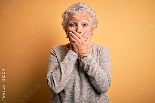 Senior beautiful woman wearing casual t-shirt standing over isolated yellow background shocked covering mouth with hands for mistake. Secret concept.