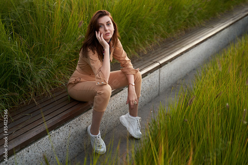 Young beautiful red haired gen z girl wearing slim fitted women beige khaki coverall overall denim jumpsuit or flight suit and white chunky platform sneakers sitting on a long park bench in high green