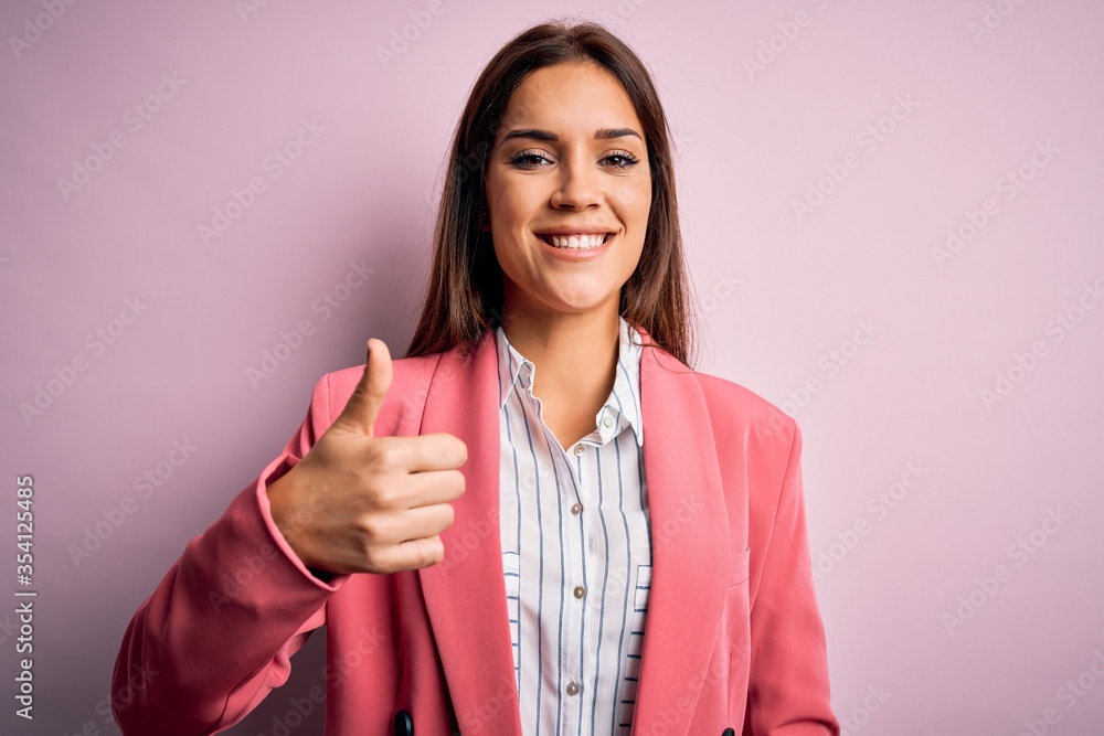 Young beautiful brunette woman wearing jacket standing over isolated pink background doing happy thumbs up gesture with hand. Approving expression looking at the camera showing success. Young beautiful brunette woman wearing jacket standing over isolated pink background doing happy thumbs up gesture with hand. Approving expression looking at the camera showing success.