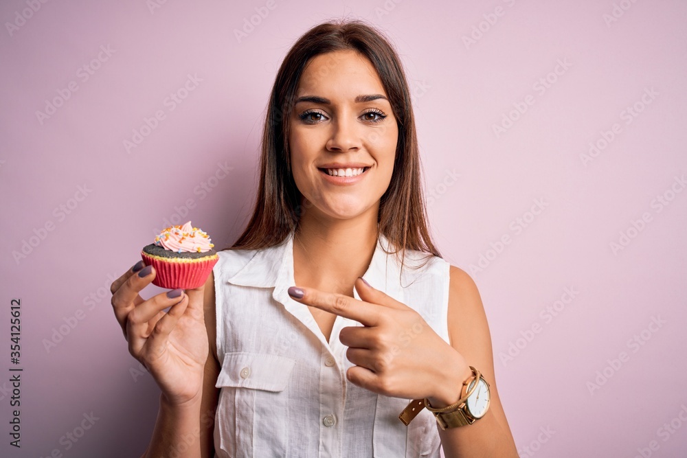 Young beautiful brunette woman eating chocolate cupcake over isolated pink background very happy pointing with hand and finger Young beautiful brunette woman eating chocolate cupcake over isolated pink background very happy pointing with hand and finger
