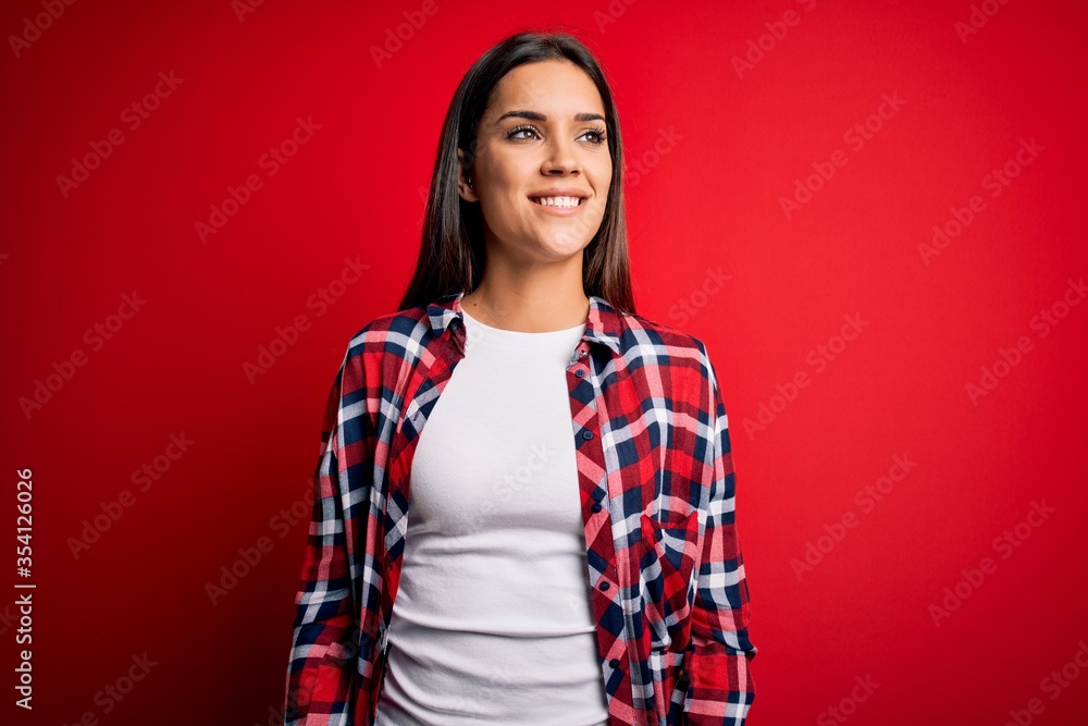 Young beautiful brunette woman wearing casual shirt standing over isolated red background looking away to side with smile on face, natural expression. Laughing confident. Young beautiful brunette woman wearing casual shirt standing over isolated red background looking away to side with smile on face, natural expression. Laughing confident.
