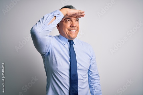 Middle age handsome grey-haired business man wearing elegant shirt and tie very happy and smiling looking far away with hand over head. Searching concept.