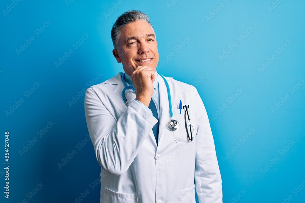 Middle age handsome grey-haired doctor man wearing coat and blue stethoscope looking confident at the camera smiling with crossed arms and hand raised on chin. Thinking positive. Middle age handsome grey-haired doctor man wearing coat and blue stethoscope looking confident at the camera smiling with crossed arms and hand raised on chin. Thinking positive.