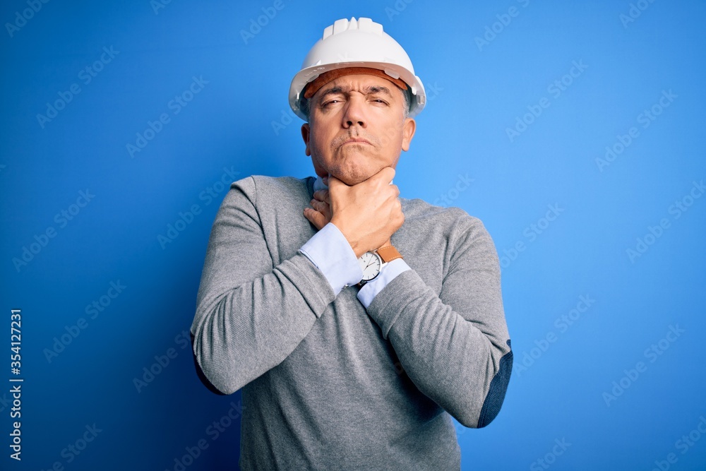 Middle age handsome grey-haired engineer man wearing safety helmet over blue background shouting and suffocate because painful strangle. Health problem. Asphyxiate and suicide concept.