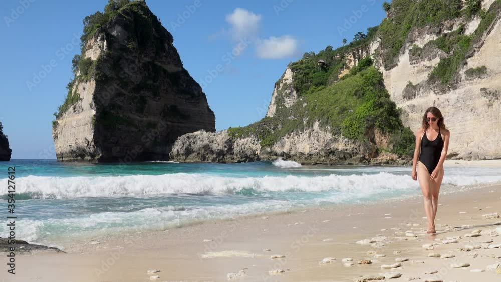 Brunette woman in black swimsuit walking on beach with sea rocks and turquoise ocean, blue sky. Atuh beach, Nusa Penida island, Bali, Indonesia. Tropical background and travel concept. Brunette woman in black swimsuit walking on beach with sea rocks and turquoise ocean, blue sky. Atuh beach, Nusa Penida island, Bali, Indonesia. Tropical background and travel concept.