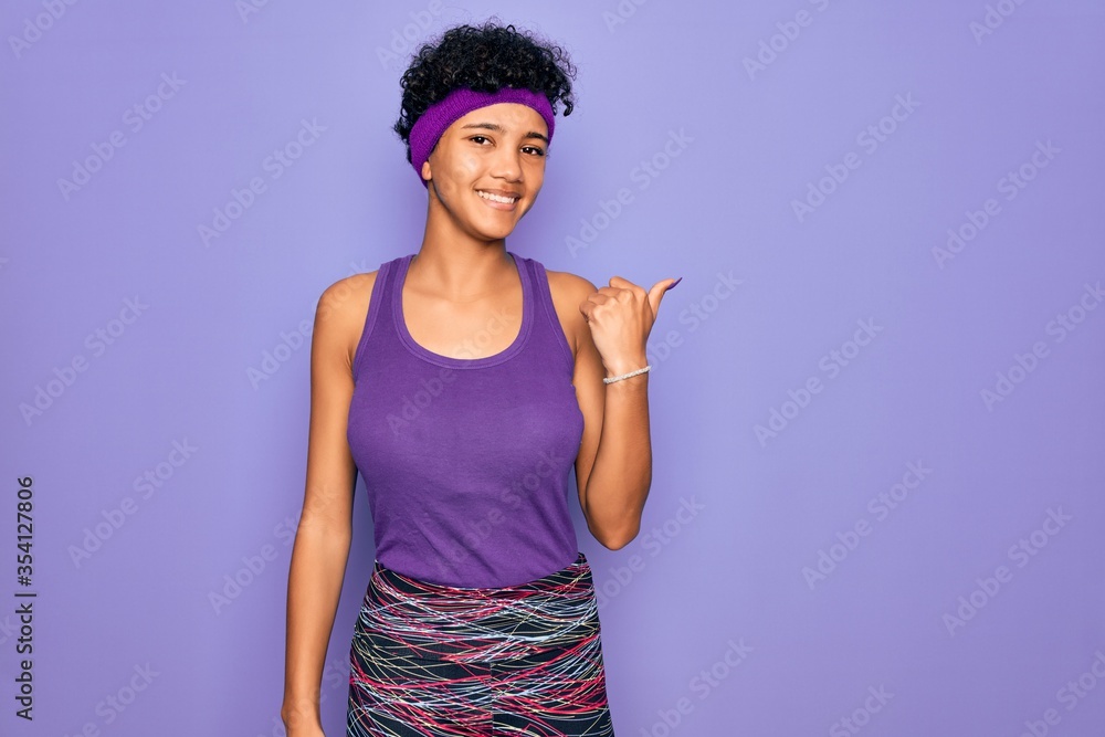 Beautiful african american afro woman wearing casual sportswear over purple background smiling with happy face looking and pointing to the side with thumb up.
