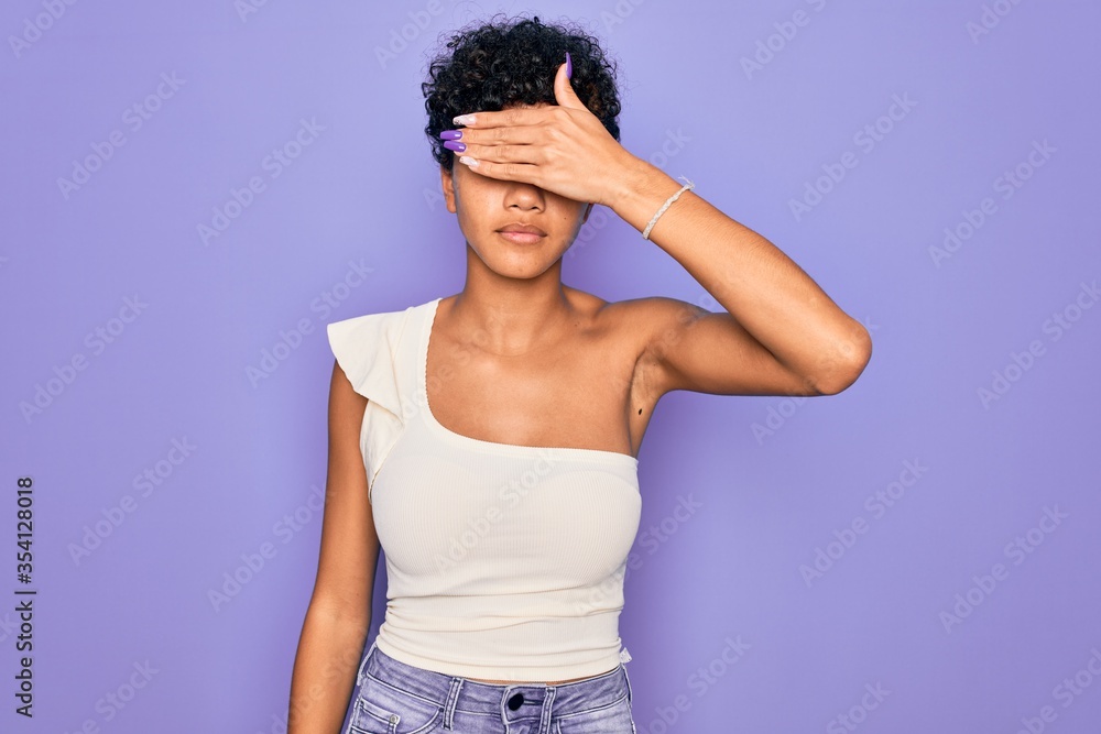 Young beautiful african american afro woman wearing casual t-shirt over purple background covering eyes with hand, looking serious and sad. Sightless, hiding and rejection concept Young beautiful african american afro woman wearing casual t-shirt over purple background covering eyes with hand, looking serious and sad. Sightless, hiding and rejection concept