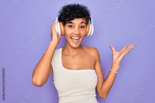 Beautiful african american woman listening to music using headphones over red background very happy and excited, winner expression celebrating victory screaming with big smile and raised hands