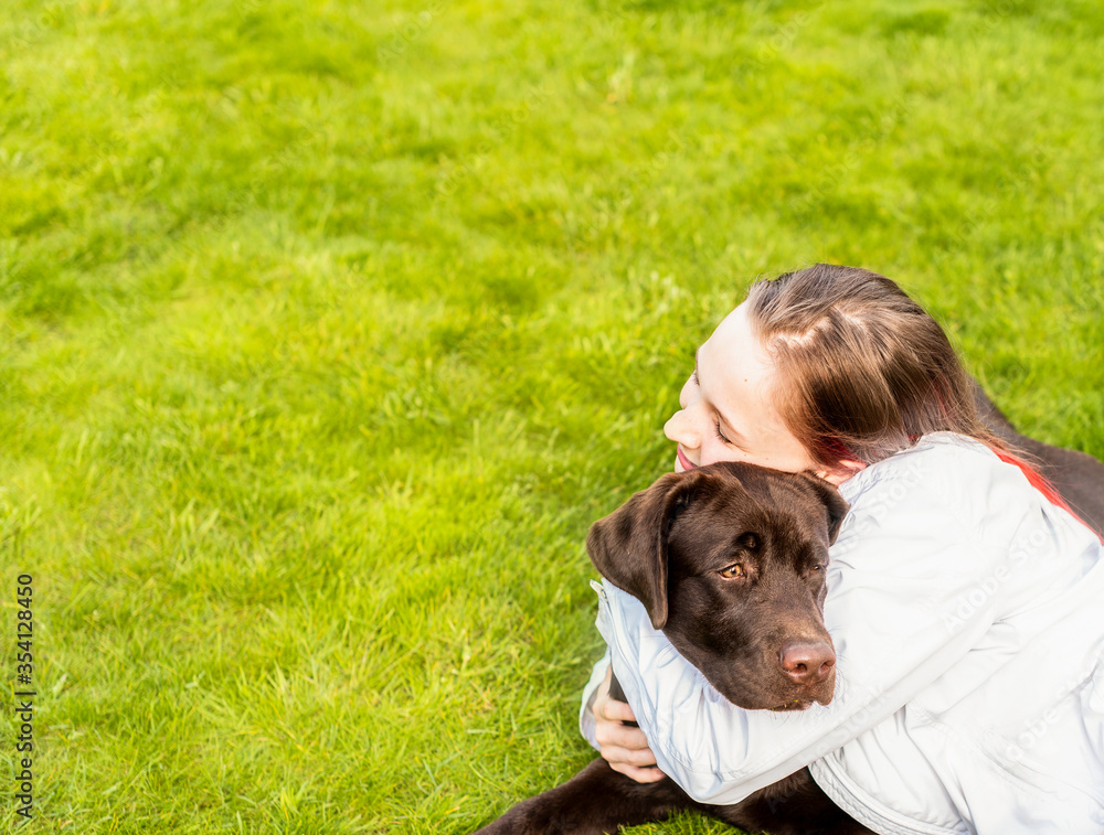 a girl in a white jacket gently and lovingly embraces a chocolate Labrador dog. They are lying in a green clearing. The girl's eyes are closed with pleasure and love. a girl in a white jacket gently and lovingly embraces a chocolate Labrador dog. They are lying in a green clearing. The girl's eyes are closed with pleasure and love.