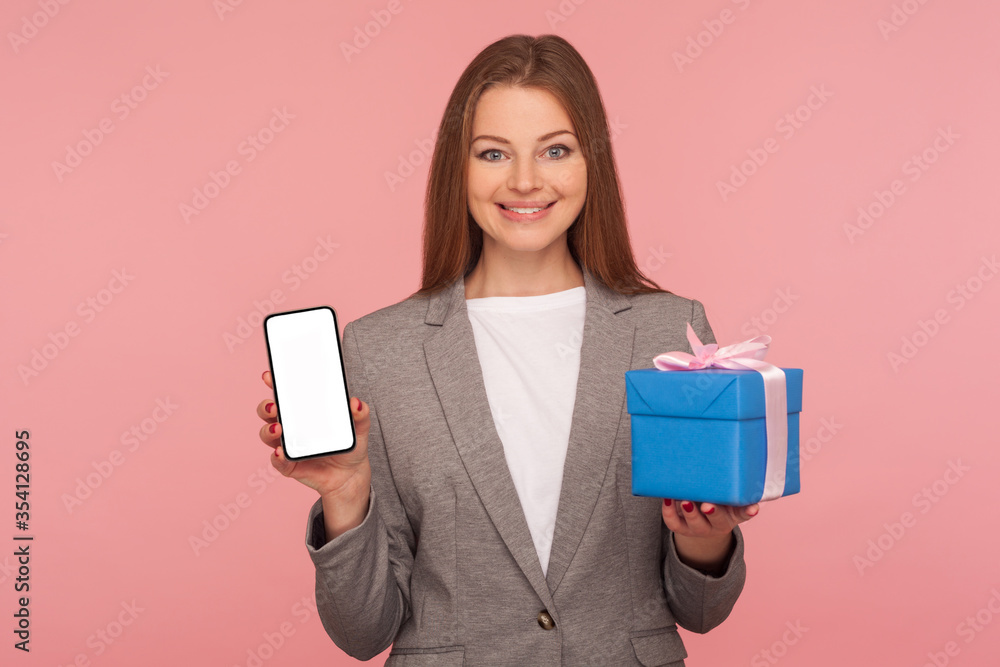 Bonus for mobile user, online shopping application. Portrait of happy elegant businesswoman in suit jacket holding cellphone and gift box, smiling to camera. studio shot isolated on pink background Bonus for mobile user, online shopping application. Portrait of happy elegant businesswoman in suit jacket holding cellphone and gift box, smiling to camera. studio shot isolated on pink background